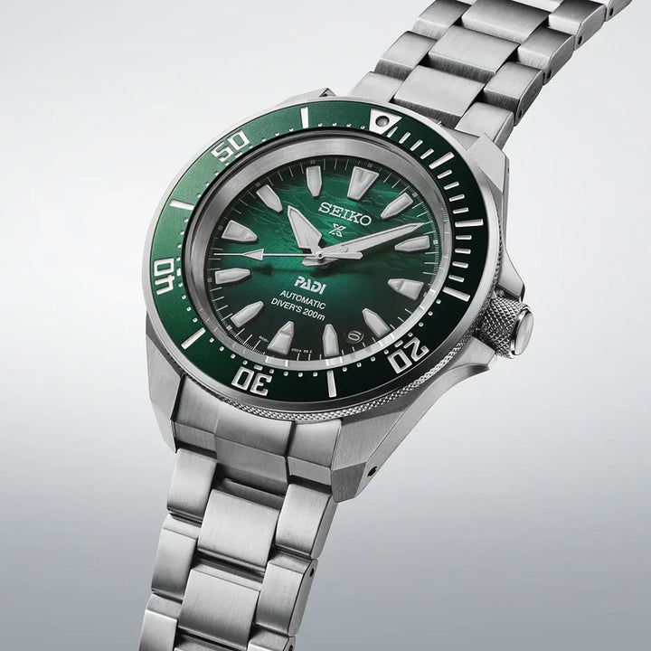 Het Seiko heren horloge SRPL53K1 heeft een groene bezel en wijzerplaat, zilveren indexen en wijzers, en een stalen band. Dit stijlvolle Seiko horloge wordt diagonaal getoond op een lichte achtergrond.