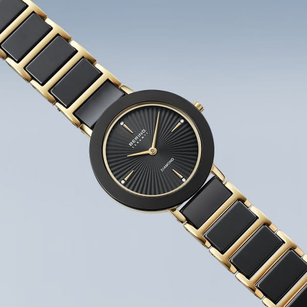 De Bering dames horloge zwarte wijzerplaat - 11429-732 heeft een zwarte en gouden metalen band, een ronde zwarte wijzerplaat met gouden markers en het Bering-logo op de wijzerplaat tegen een lichtblauwe achtergrond met kleurverloop.