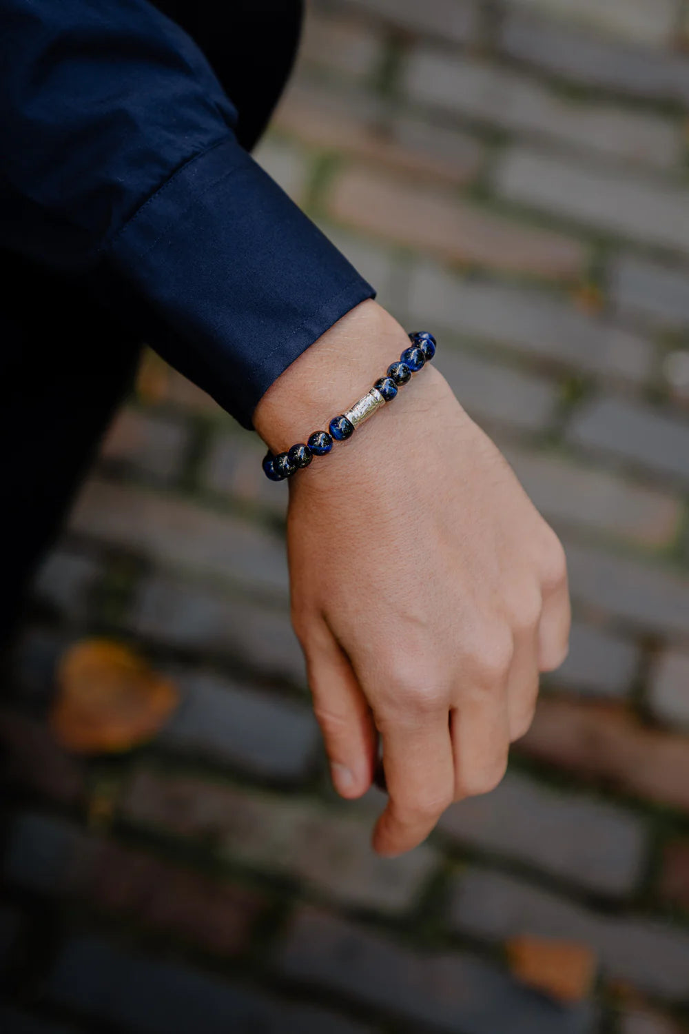 Een hand versierd met de AZE Jewels Kralen armband heren - Mount Blue Maine - 8mm, toont een blauwe en zilveren kralenarmband tegen een geplaveid pad bezaaid met bruine bladeren, wat doet denken aan een herfstwandeling. Marineblauwe overhemdmouw zichtbaar.