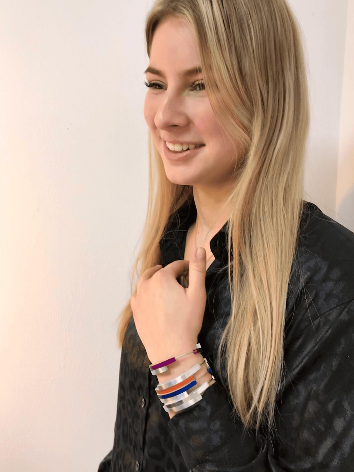 Een vrouw met lang blond haar lacht in een shirt met patroon, houdt haar kraag vast en toont verschillende levendige CLIC Creations-armbanden, waaronder de Armband Vier bogen Zwart A32Z. De achtergrond is een effen witte muur.
