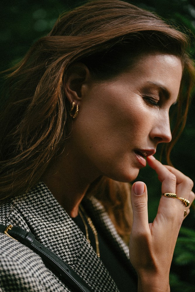 Een vrouw met golvend bruin haar draagt een houndstooth blazer, Excellent Jewelry Geelgouden Oorringen met Gestreept Patroon (14 karaat, Van der Kooij), en een gouden ring tikt nadenkend haar lippen buiten tussen het groen.