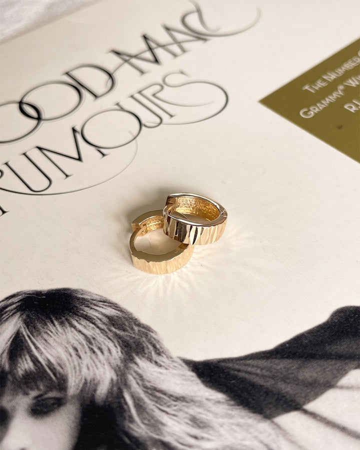 Twee Geelgouden Oorringen met Bewerkte Voorzijde van Excellent Jewelry, die subtiele luxe uitstralen, rusten op een Fleetwood Mac Rumours albumhoes met de gedeeltelijke titel en een zwart-wit afbeelding van een langharige persoon.