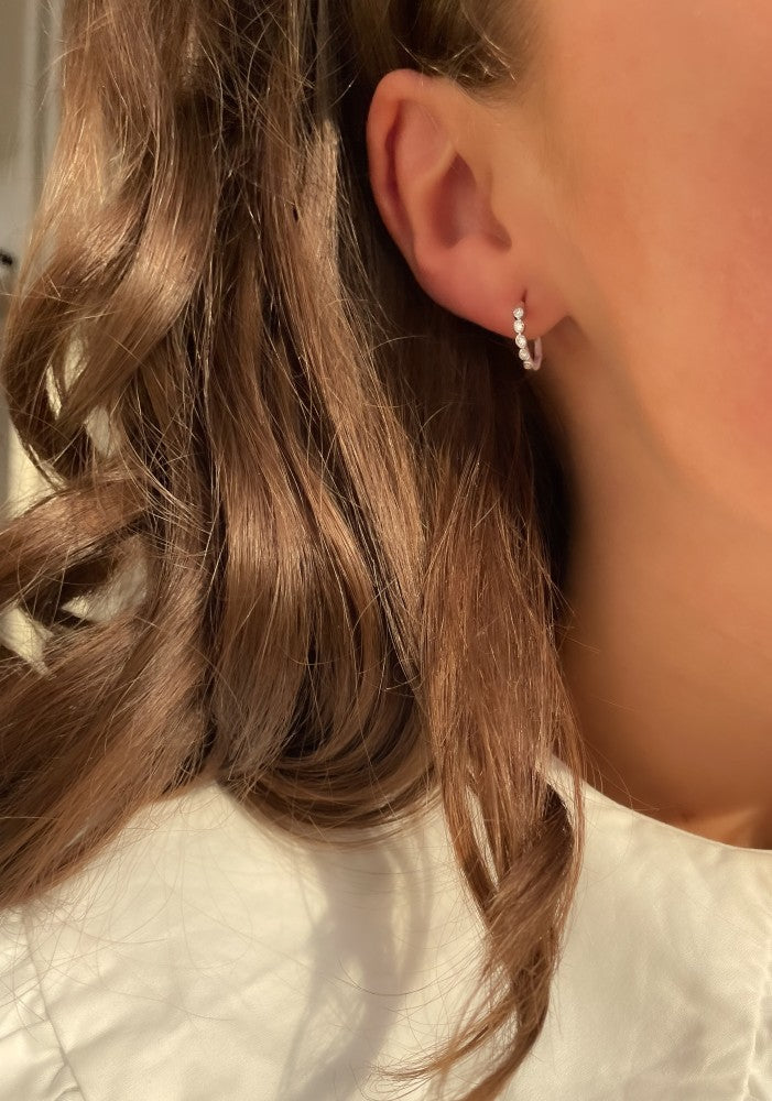 Close-up van het oor van een vrouw met Oorringen Luna 0,20 crt. witgoud van Aurore Jewelry, geaccentueerd met kleine pareltjes. Haar bruine haar valt in losse golven en ze is gekleed in een licht shirt, badend in warm zonlicht.