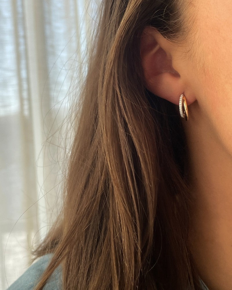 Close-up van een vrouwenoor met lang bruin haar, dat Excellent Jewelry 14 Karaat Bicolor Gouden Oorstekers met Diamant - 0,18 crt Briljant draagt. Lichte, transparante gordijnen en zacht natuurlijk licht creëren een zachte sfeer op de achtergrond.