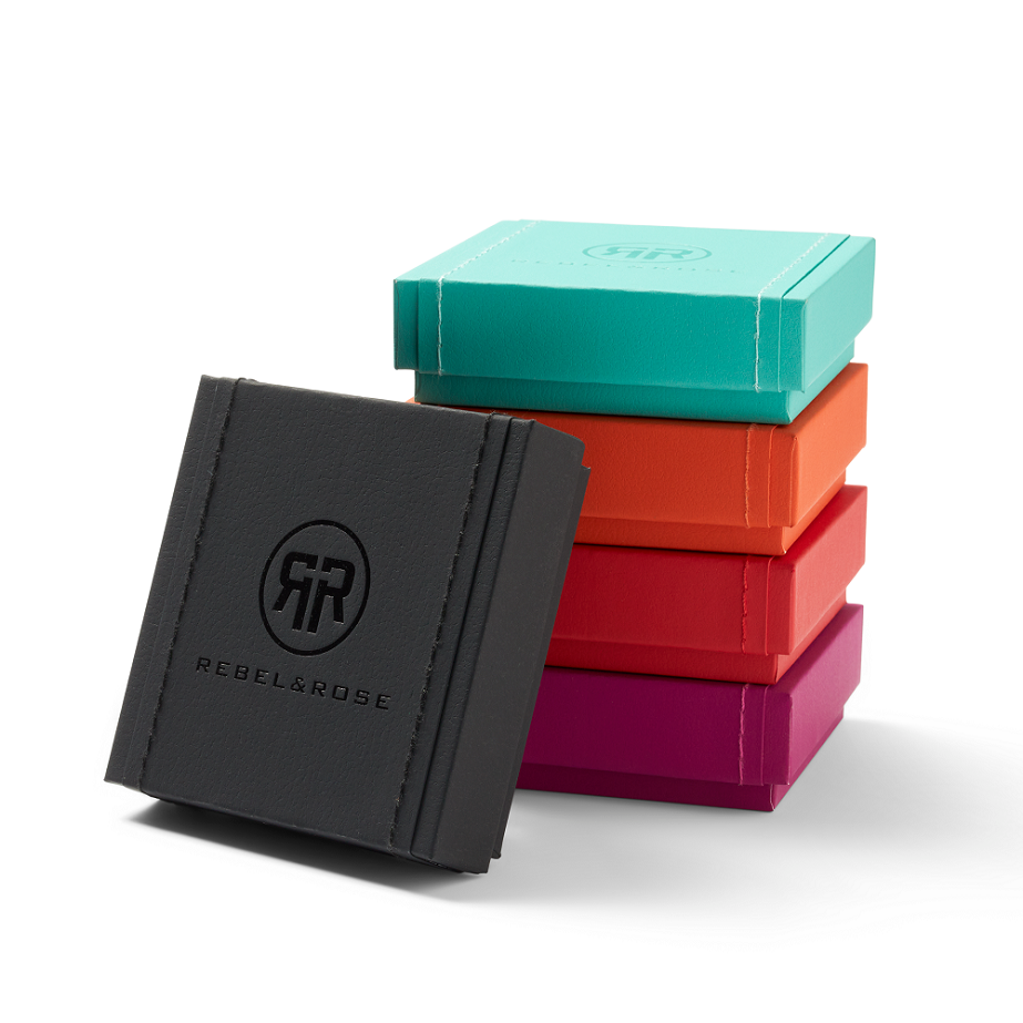 Vijf kleurrijke vierkante sieradendoosjes zijn gestapeld, waarbij het zwarte doosje aan de voorkant het Rebel & Rose logo toont. Ideaal om een luxe sieraad cadeau te doen, zoals de Mix Tiger Lapis - 8mm - Heren Kralen Armband van Rebel & Rose. Effen witte achtergrond.