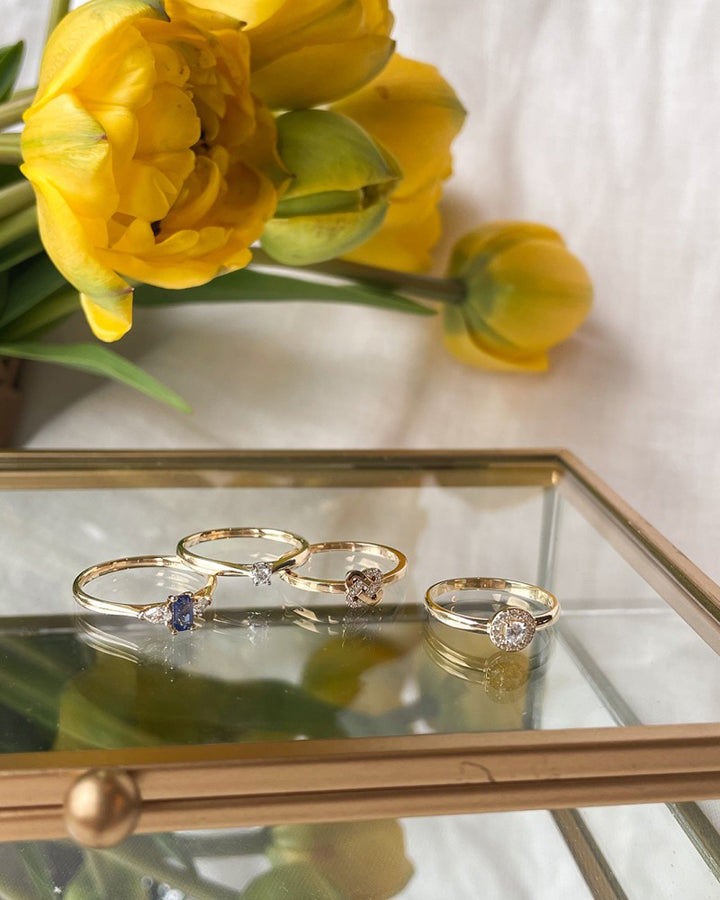 Vier 14kt gouden ringen, waaronder de Klassieke 14kt Gouden Solitair Ring met 0.06ct Diamant van Excellent Jewelry, worden tentoongesteld op een glazen sieradendoosje. Gele tulpen lichten de achtergrond op op een licht oppervlak.