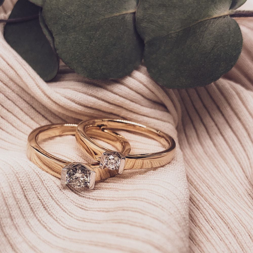 Twee gouden ringen, waaronder de 14kt Gouden Ring met 0,10ct Briljant Diamant van Excellent Jewelry, rusten op zachte geribbelde beige stof met groene eucalyptusbladeren erboven, waardoor een elegant en romantisch tafereel ontstaat.