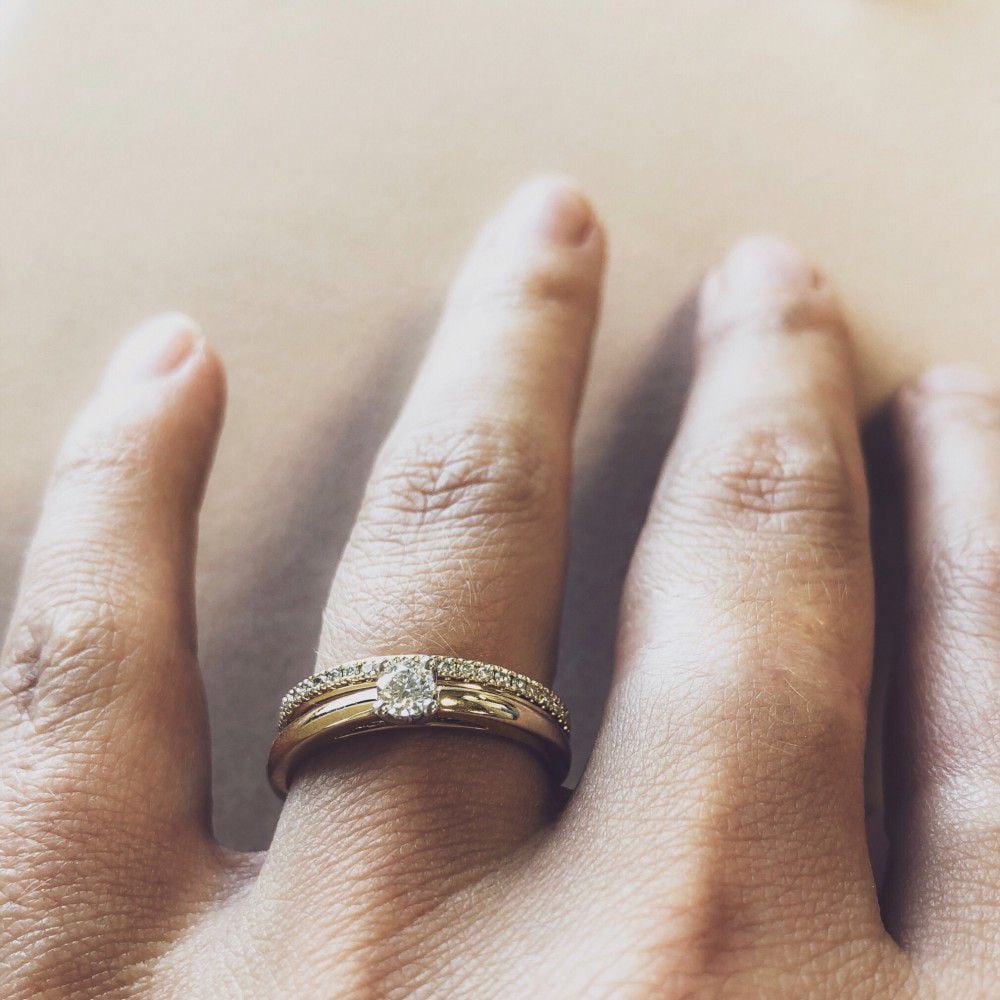 Een close-up toont een hand met een 14 karaat gouden band en de Excellent Jewelry Bicolor Wit- en Geelgouden Solitair met Briljant 0.19 crt., beide aan de ringvinger, met een zacht vervaagde neutrale achtergrond.
