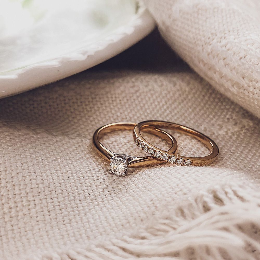 Twee Elegante Bicolor Gouden Ringen van Excellent Jewelry liggen op beige stof; één ring heeft een enkele diamant, de andere is bezet met briljanten van 0.19 crt.