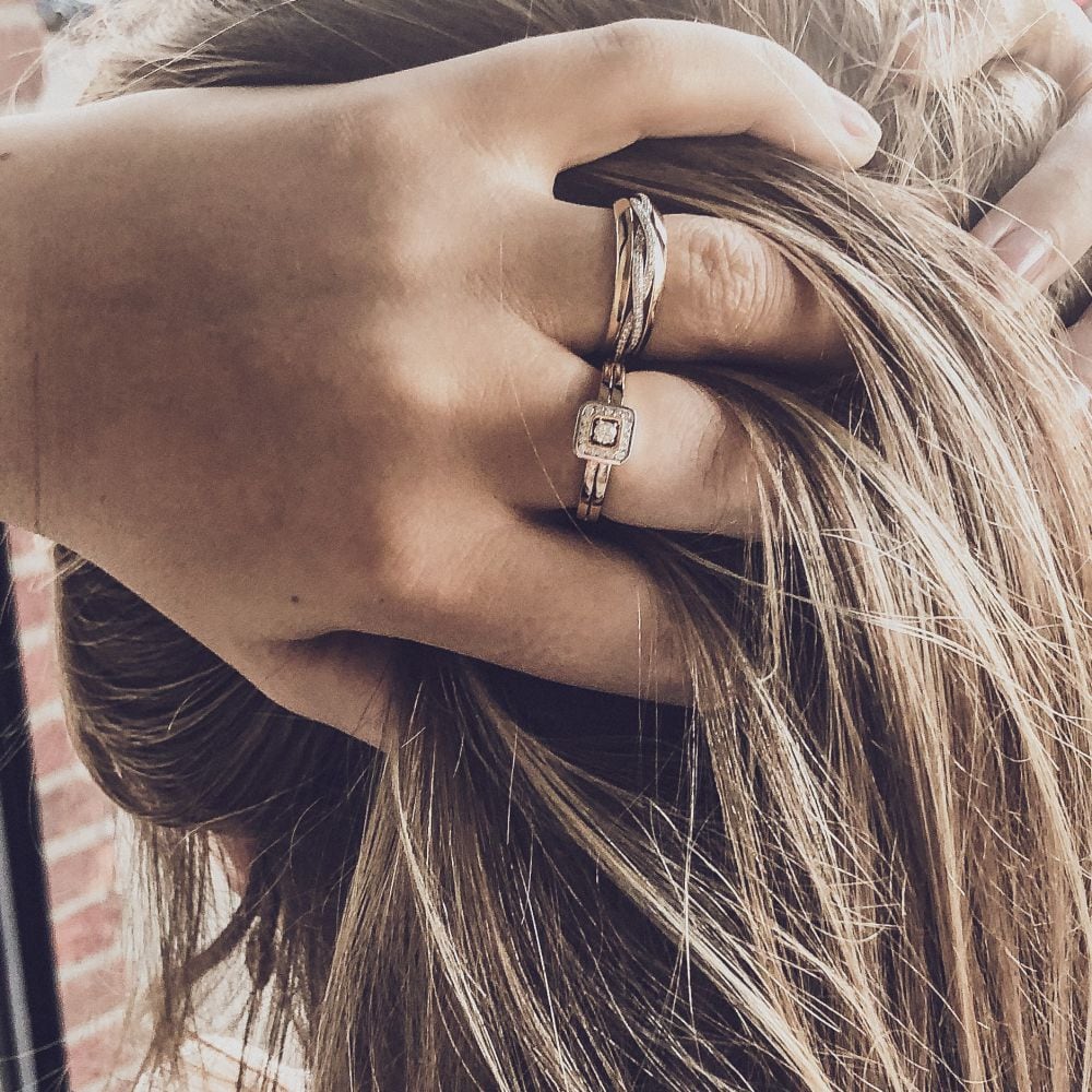 Een close-up toont een hand met twee Bicolor Geel- en Witgouden Ringen met Diamant - 0,22 ct van Excellent Jewelry, die zachtjes iemands hoofd vasthoudt, vingers in lang lichtbruin haar tegen een zacht vervaagde achtergrond.