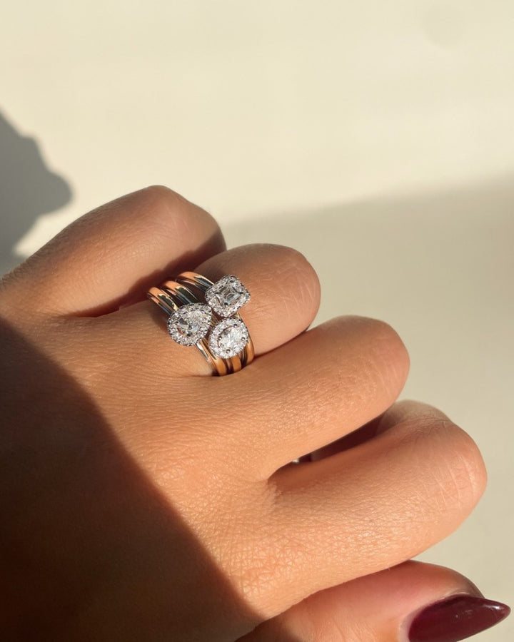 Een hand met gemanicuurde nagels toont drie gouden ringen, waaronder de 14k Bicolor Gold Ring van Excellent Jewelry met een ovaal diamanten hart in een entourage zetting, die allemaal schitterend schitteren in natuurlijk zonlicht.