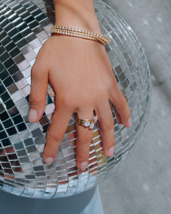 Een hand met gemanicuurde nagels draagt de 14k Bicolor Gouden Ring met Ovale Diamant Kern van Excellent Jewelry in een entourage zetting, rustend op een glimmende discobal die kleine lichtvierkantjes reflecteert.
