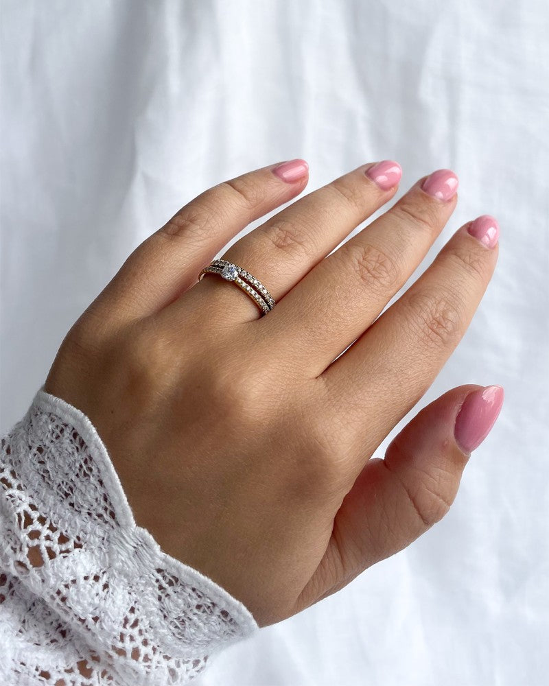 Een hand met lichtroze nagels en een kanten mouwtje toont de Elegante bicolor solitair ring met briljant geslepen diamant 0.35 crt. van Excellent Jewelry, zacht glinsterend tegen een witte achtergrond.