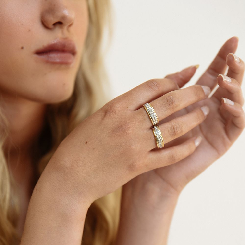 Een close-up van een vrouw met een lichte huid en blond haar die twee fonkelende damesringen draagt, waaronder de Excellent Jewelry Elegante damesring met geelgouden band en 0,54ct briljant diamant. Haar natuurlijke nagels en de zacht vervaagde achtergrond maken de look compleet.