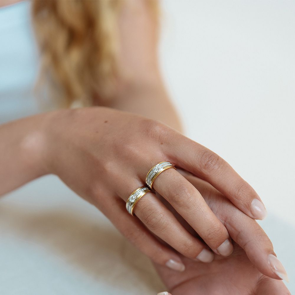 Een close-up van een hand met gemanicuurde nagels die twee Elegante damesringen van Excellent Jewelry draagt, geelgouden band met 0,54ct briljant diamant, rustend op een licht oppervlak. Lang golvend blond haar verschijnt op de achtergrond.