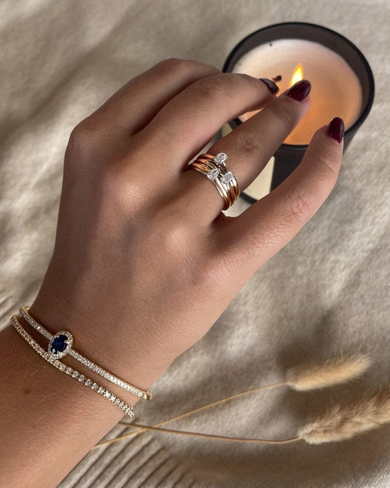 Een hand met bordeauxrode nagels draagt gestapelde ringen, waaronder de Excellent Jewelry 14k Bicolor Gold Solitaire Ring met Ovale Diamant van 0,19ct, plus twee diamanten armbanden, waarvan één met een blauwe edelsteen, terwijl ze een brandende kaars op beige stof vasthoudt. Gedroogd gras is dichtbij.