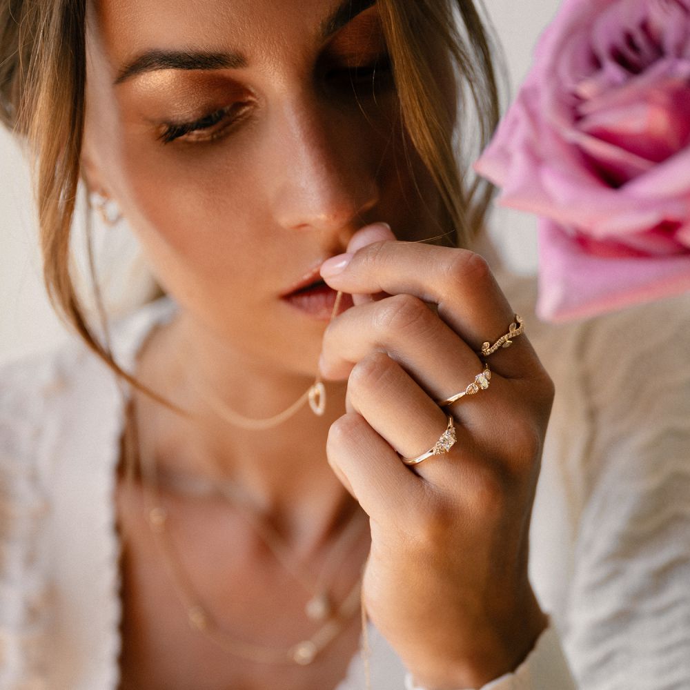 Een vrouw met lichtbruin haar draagt gelaagde gouden kettingen en de Vintage Driesteens Geelgouden Ring met Ovale Diamant en Peer Geslepen Zijstenen (14K, 0,35 ct, H/SI-1) van Excellent Jewelry, starend naar beneden met een roze roos naast haar gezicht.