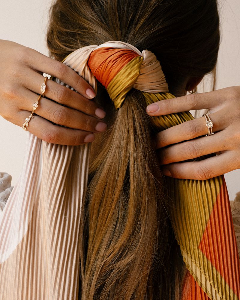 Een vrouw bindt haar bruine haar vast met een geplooide, veelkleurige sjaal in oranje, geel en crème. Op haar handen zitten Excellent Jewelry 14 karaat Vintage Driesteens Geelgouden Ringen met ovale diamant en peer geslepen zijstenen, 0,35 ct.