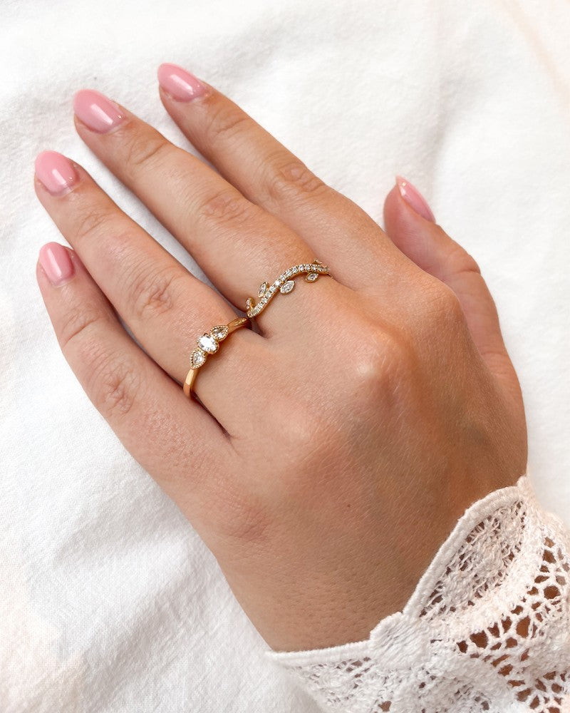 Een hand met roze gepolijste nagels draagt de Vintage Driesteens Geelgouden Ring met Ovale Diamant en Peer Geslepen Zijstenen - 14 karaat, 0,35 ct van Excellent Jewelry en een andere fijne diamanten ring aan de ringvinger, tegen witte stof met een kanten mouwtje.