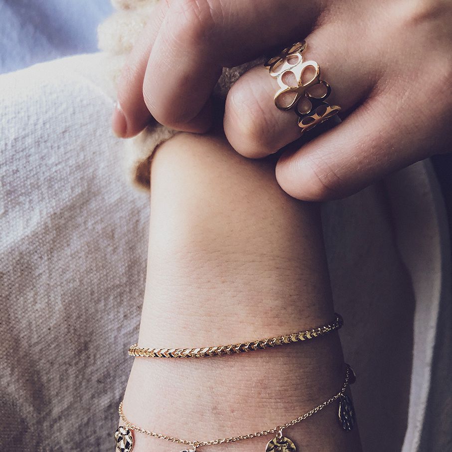 Twee handen tonen delicate gouden sieraden; de ene draagt de Excellent Jewelry Geelgouden Damesring met Opengewerkt Bloemenpatroon in 14 karaat goud, terwijl de andere dunne gouden armbanden draagt. Een zachte achtergrond versterkt hun elegante uitstraling.