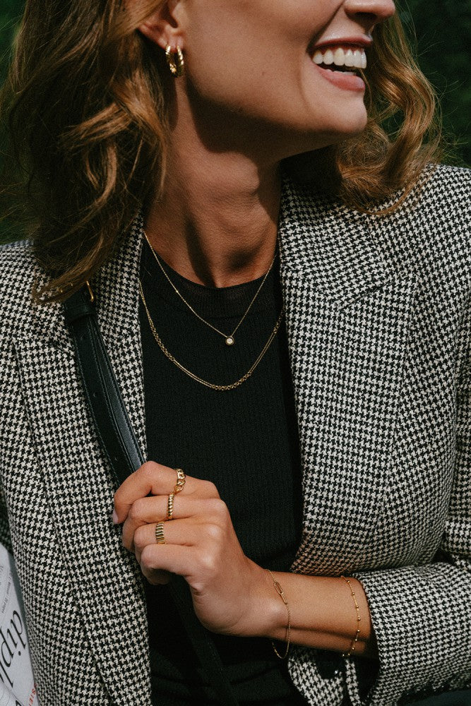 Een vrouw in een zwarte top en houndstooth blazer glimlacht, draagt gouden hoepel oorbellen, gelaagde kettingen, een delicate armband, en de Excellent Jewelry Geelgouden Gedraaide Bandring - 14kt goud, 4,3mm breed. Ze houdt een zwarte band vast; groen staat achter haar.