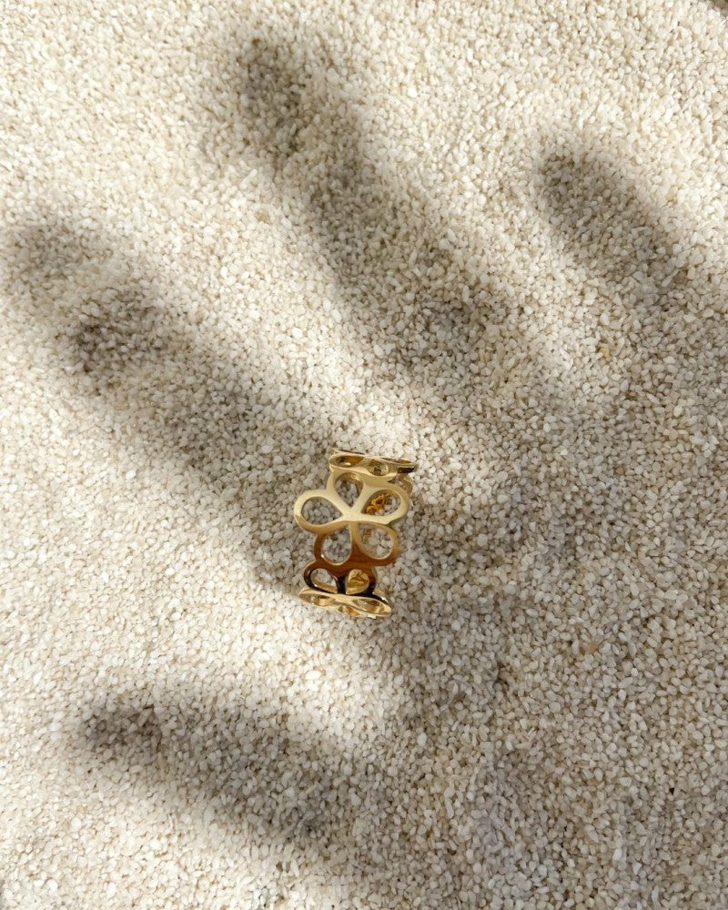 De Geelgouden Damesring met Opengewerkt Bloemenpatroon van Excellent Jewelry, vervaardigd uit 14 karaat goud, ligt op licht zand; haar schaduw versmolten met die van een uitgestrekte hand die ernaar reikt.