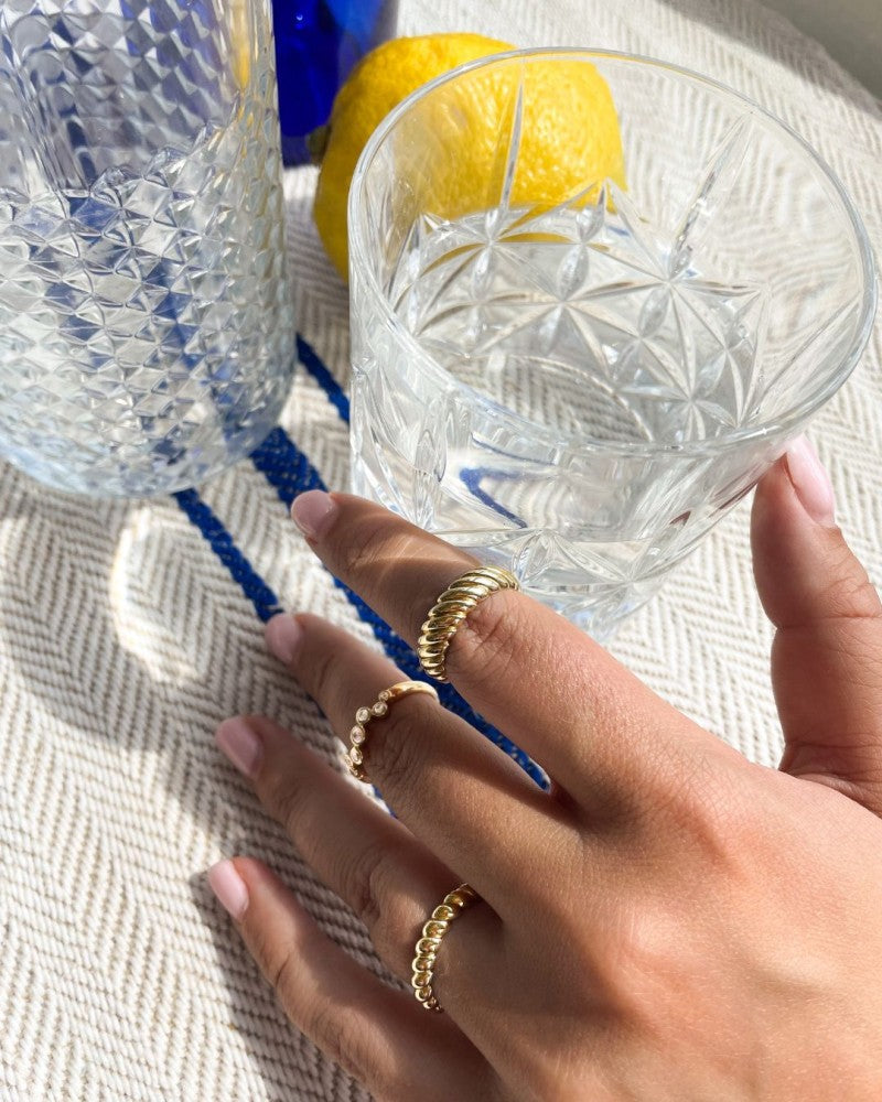 Een hand die de Geelgouden Gedraaide Bandring van Excellent Jewelry draagt - 14kt goud, 4,3 mm ras - houdt een glazen beker met patroon vast; een hele citroen en een ander glas rusten vlakbij terwijl het zonlicht zachte schaduwen werpt over het gestructureerde oppervlak.