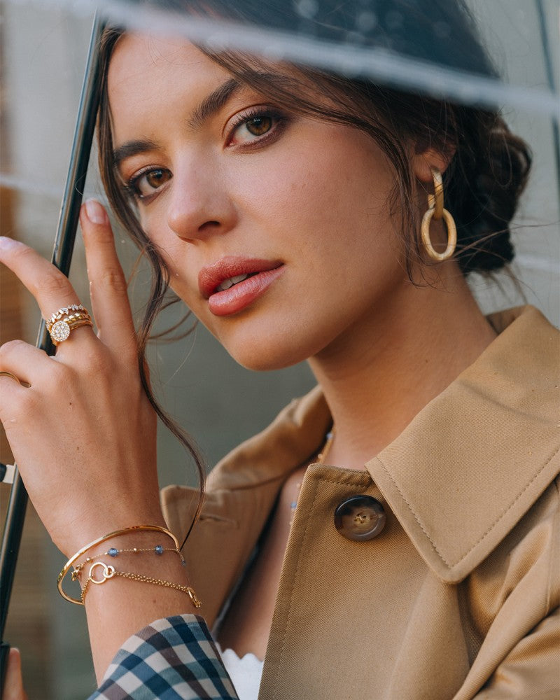 Een vrouw met bruin haar in een los kapsel houdt een doorzichtige paraplu vast, draagt een beige trenchcoat, gouden hoepeloorbellen en de Excellent Jewelry Geelgouden Gedraaide Bandring - 14kt goud, 4,3 mm ras, en kijkt nadenkend naar de camera.