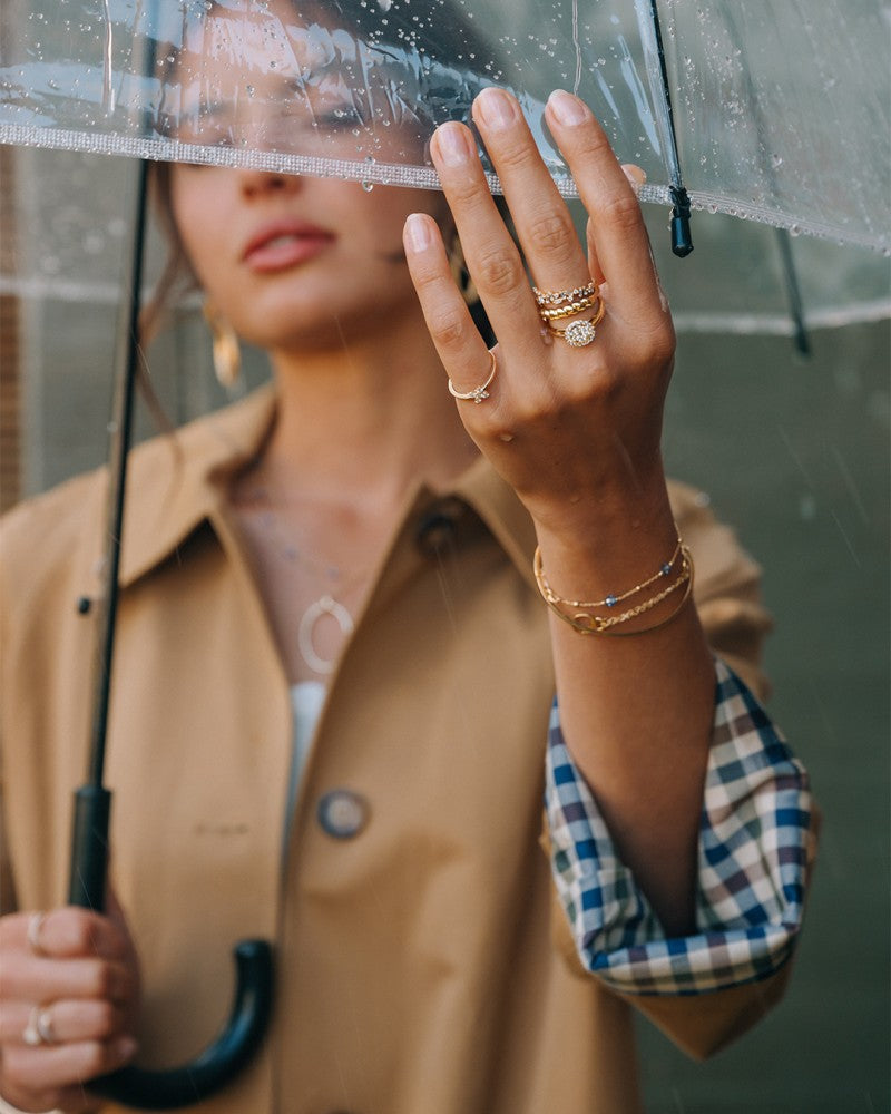 Een vrouw in een tan trenchcoat en wit topje houdt een doorzichtige paraplu vast in de regen, waardoor haar Excellent Jewelry Geelgouden Gedraaide Bandring - 14kt goud, 4,3 mm breed, 1,8 gram. Haar gezicht is gedeeltelijk onscherp.