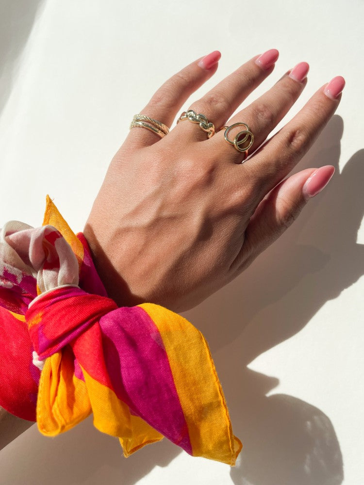 Een hand met roze gemanicuurde nagels showt de Excellent Jewelry Geelgouden Damesring met Speels Design - 14 Karaat Goud, met een kleurrijke sjaal om de pols die een schaduw werpt op een witte achtergrond.