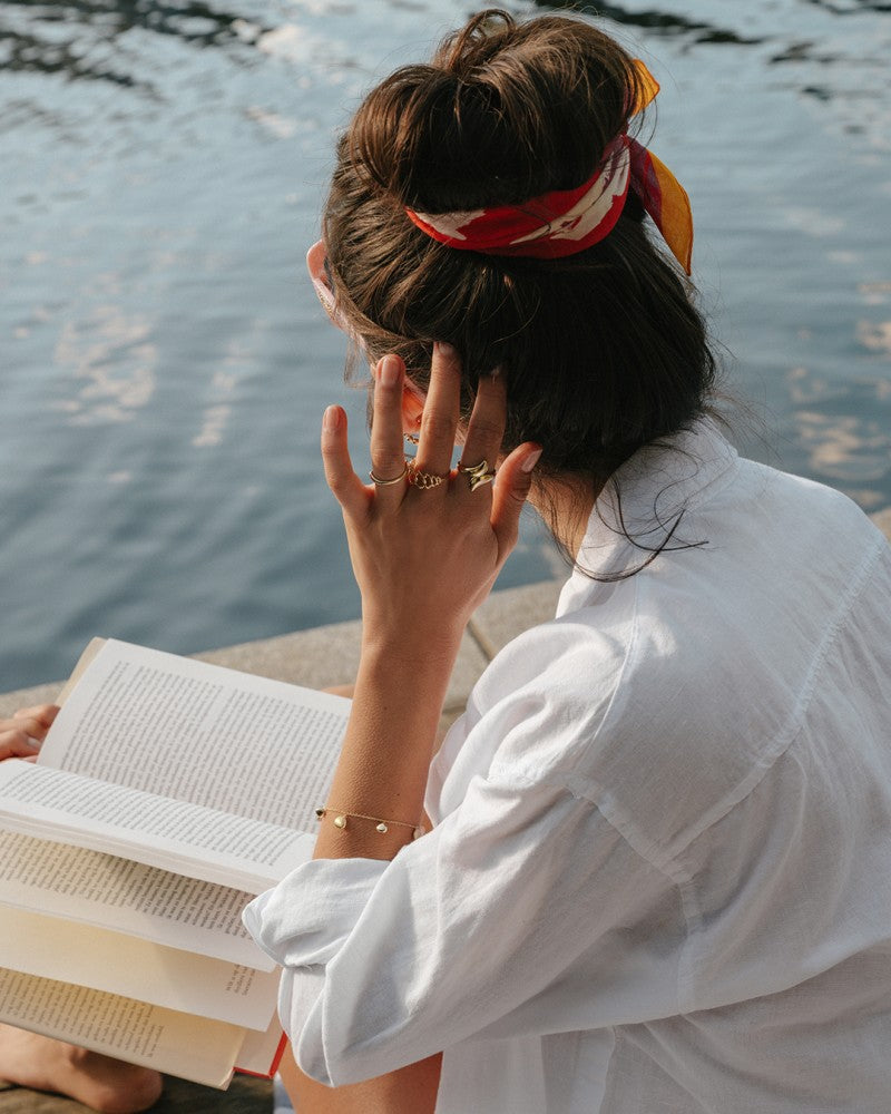 Een vrouw met een rood gedessineerde sjaal zit aan het water, draagt een wit shirt en de Opvallende Geelgouden Ring met Speels Design van Excellent Jewelry, leest een boek en strijkt door haar haar.