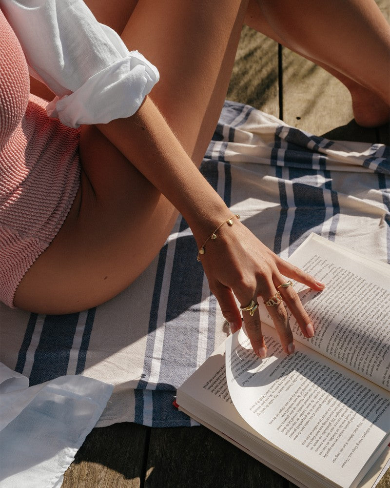 Een persoon die gouden sieraden draagt, waaronder de Opvallende Geelgouden Ring met Speels Design van Excellent Jewelry, leest buiten een boek op een geruite deken. Zonlicht benadrukt hun blote benen en opgerolde witte overhemdmouw.