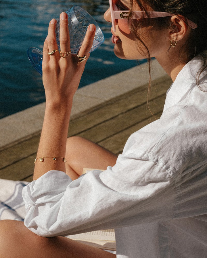 Een vrouw in een wit shirt en een zonnebril ontspant aan het water, terwijl ze een blauw glas vasthoudt. Ze draagt een Excellent Jewelry Opvallende Geelgouden Ring met Speels Design - Een Vleugje Verfijnde Stijl om haar vinger terwijl zonlicht haar blik benadrukt.