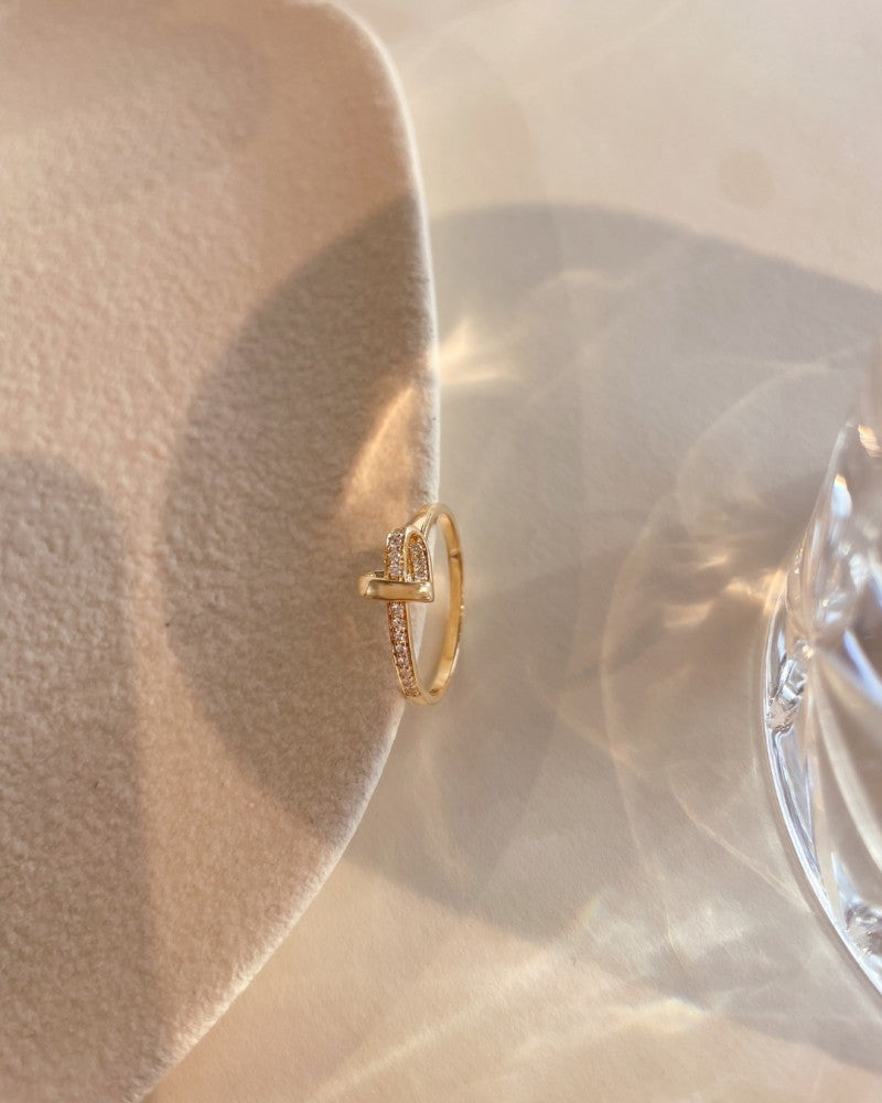 De Geelgouden Hart Ring met 0,11ct diamanten van Excellent Jewelry rust op een zachtbeige gestructureerd oppervlak naast een helder glas dat reflecties op de tafel werpt.