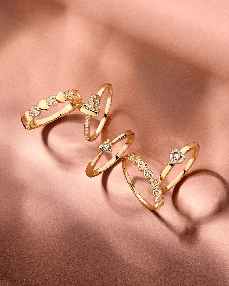 De Geelgouden Hart Ring met Diamanten, 0.11ct van Excellent Jewelry schittert tussen vijf gouden ringen op perzikkleurig satijn, die delicate schaduwen werpen en het ingewikkelde ontwerp benadrukken.