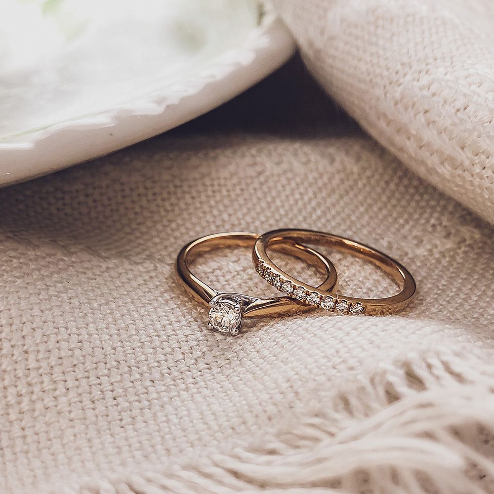 Op beige geweven stof liggen Gouden Oorknoppen met Diamant (0,12 karaat, 14 karaat geel goud, briljante slijpvorm) van Excellent Jewelry; op de achtergrond is een witte keramieken schaal deels zichtbaar.