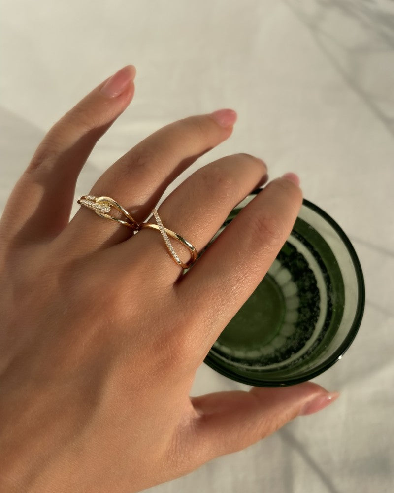 Een gemanicuurde hand houdt een groen glas vast en draagt twee gouden ringen van Excellent Jewelry - de ene is de "Ring geelgoud briljant 0.12 crt." en de andere heeft een delicaat slangenontwerp. Zonlicht werpt zachte schaduwen op een wit oppervlak.