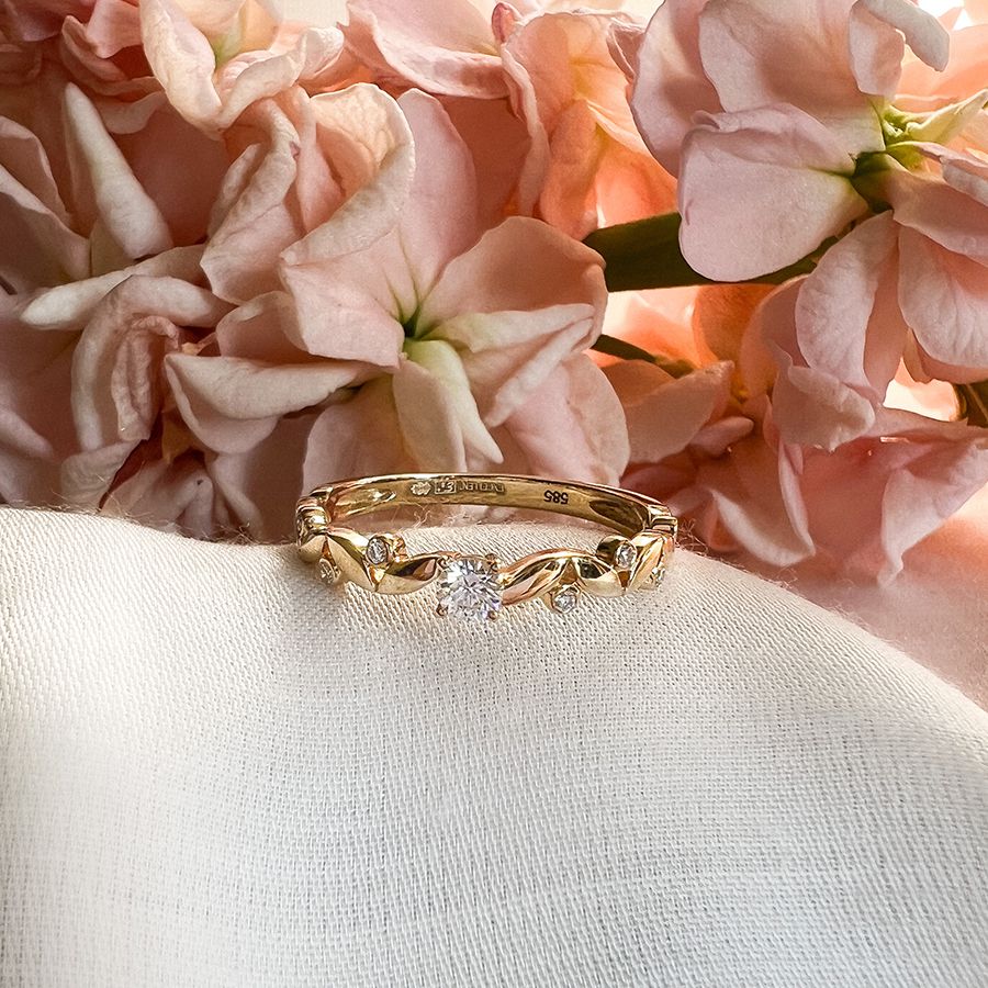 De Elegante Geelgouden Ring met 0,16 krt Briljant Diamant van Excellent Jewelry roest op witte stof, omringd door zachte roze bloemen. Subtiele luxe voor dames straalt uit dit verfijnde sieraad.