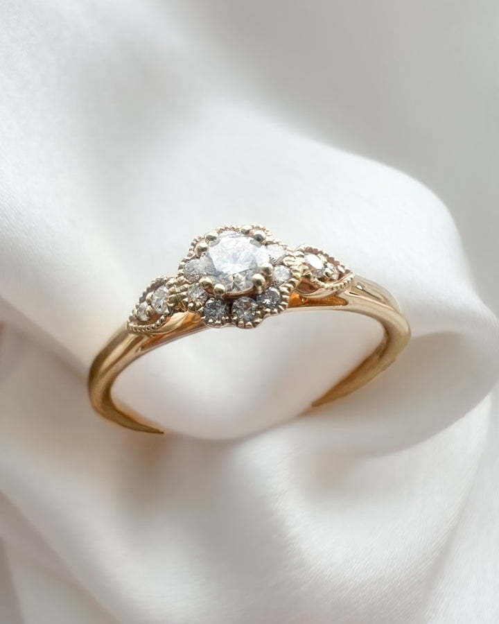 De Verfijnde Geelgouden Ring met Briljant Geslepen Middensteen 0.21 crt. van Excellent Jewelry schittert met een centrale briljant, omlijst door kleinere diamanten en gepresenteerd op witte stof.