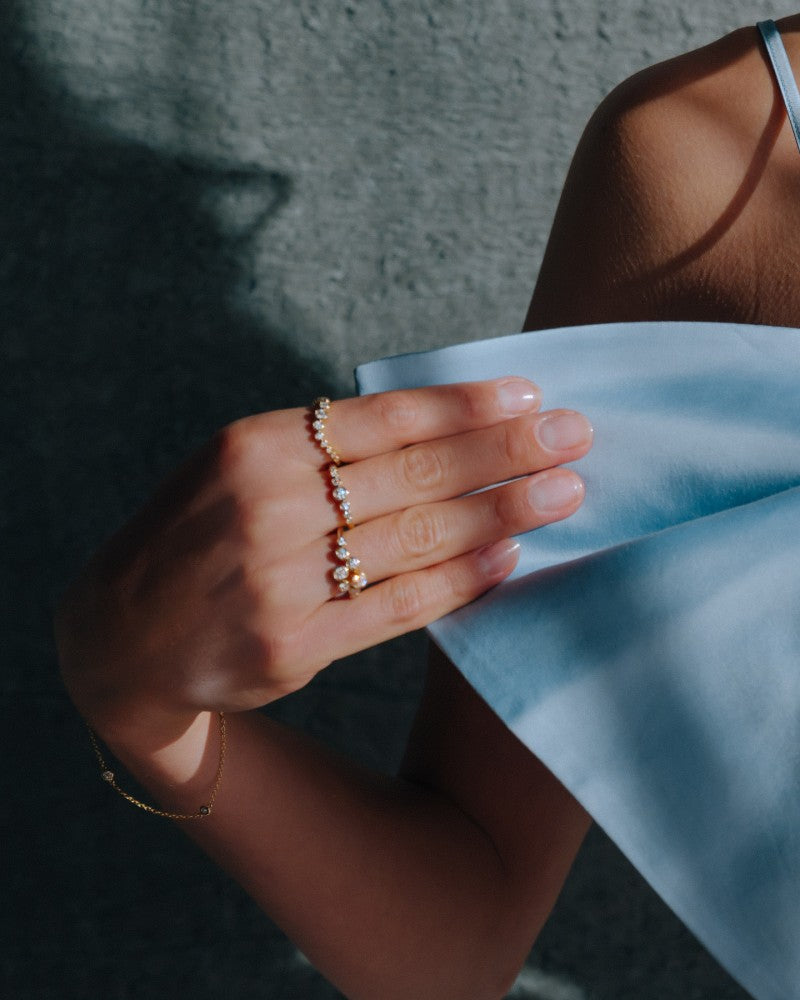 Een persoon in een lichtblauwe off-shoulder top houdt de stof zachtjes vast en showt de Playful 14k Gouden Ring met 0,37ct Diamanten uit de 'Bubbles' Collectie van Excellent Jewelry om haar vinger, gecombineerd met een dunne gouden armband en parels.