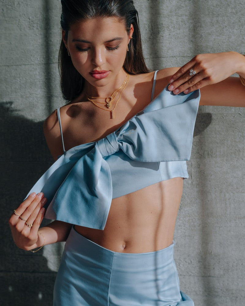 Een vrouw met een lichtblauwe one-shoulder crop top met een grote strik en bijpassende rok staat tegen een grijze muur met textuur, naar beneden kijkend terwijl ze haar top verstelt. Ze draagt de 'Bubbles' 14k Geelgouden Ring van Excellent Jewelry (0,37ct diamanten).