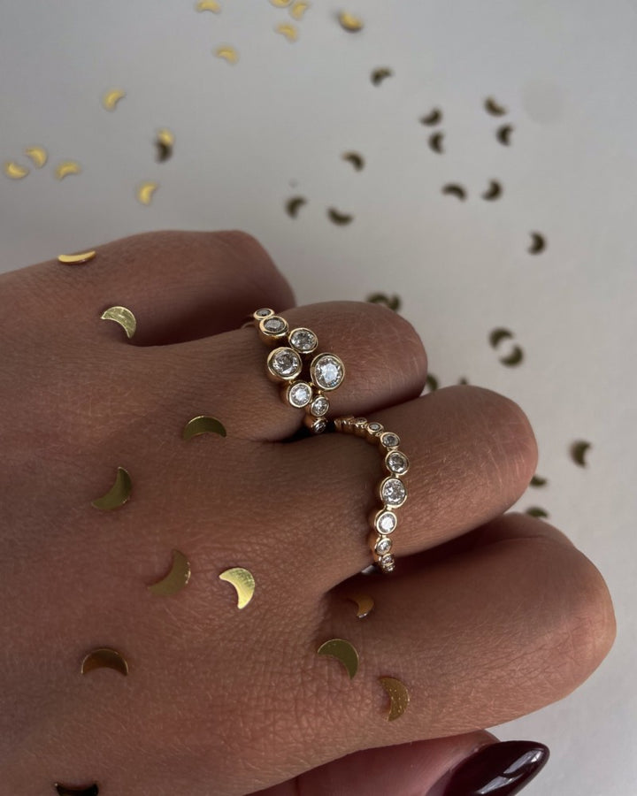 Een hand versierd met gouden confetti draagt de Speelse 14k Gouden Ring met 0,37ct diamanten uit de 'Bubbles' Collectie van Excellent Jewelry. De golvende geelgouden band schittert tegen een lichte achtergrond die bezaaid is met nog meer confetti.