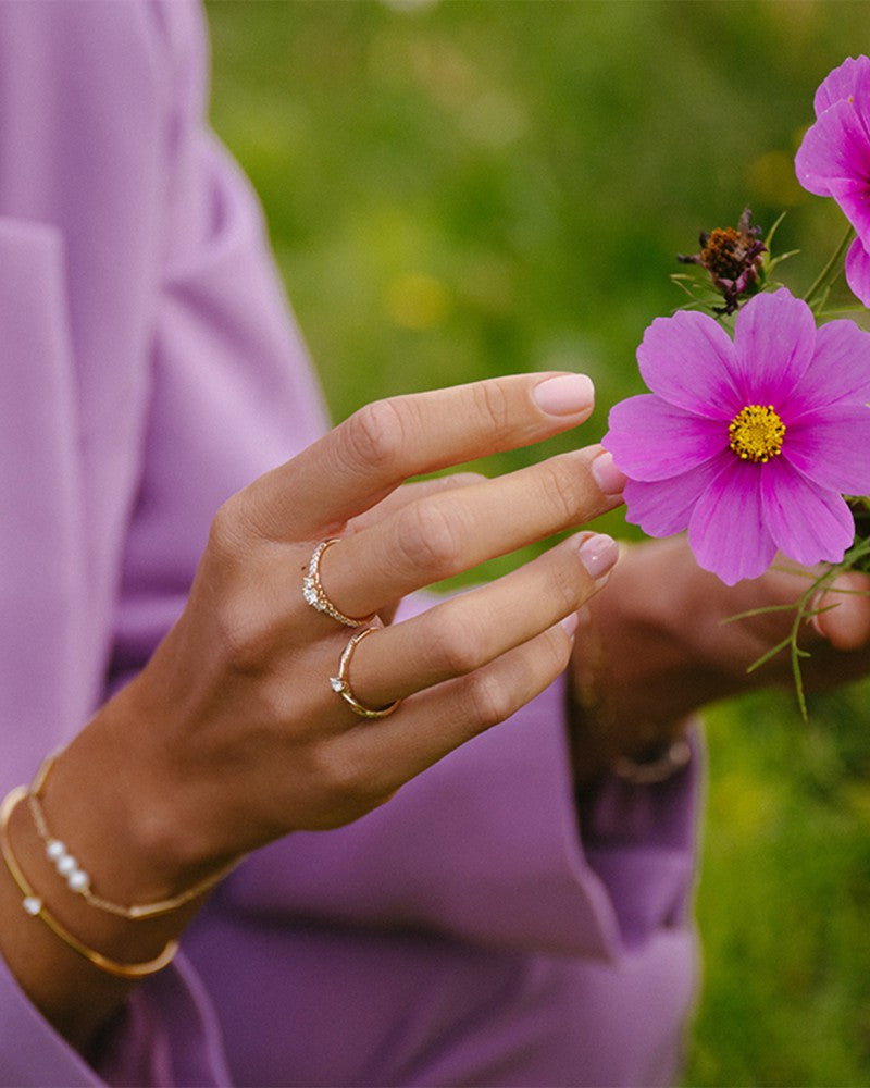 Met gouden armbanden, een diamanten ring en de Excellent Jewelry Geelgouden Driesteens Ring met Extra Diamanten in de Scheen voor Maximale Schittering, raakt een persoon in een lavendelkleurig topje voorzichtig een roze bloem aan met vaag groen gras op de achtergrond.