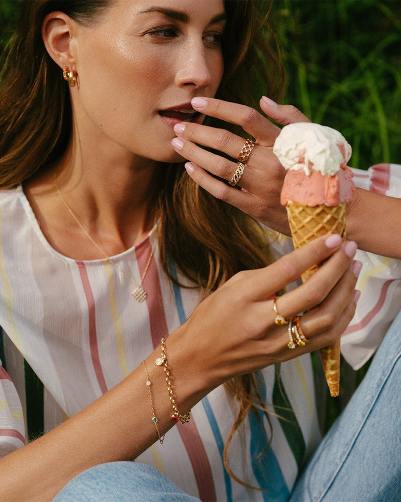 Een vrouw met gouden sieraden en een Excellent Jewelry Prachtige Geelgouden Ring in Geweven Design, gezet met fonkelende diamanten 0.46 crt, houdt bedachtzaam een ijsje met twee bolletjes vast terwijl ze buiten op het gras zit.