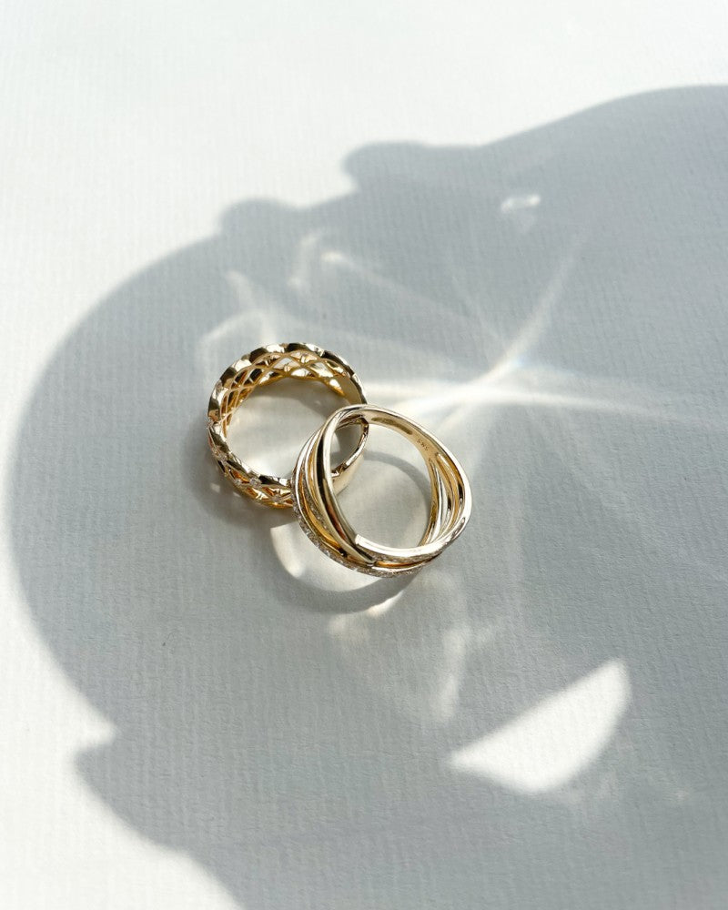 Twee gouden ringen liggen op een wit gestructureerd oppervlak: de ene is Excellent Jewelry's Prachtige Geelgouden Ring in Geweven Design, met 0,46 crt. fonkelende diamanten; de andere heeft drie in elkaar grijpende banden. Zachte schaduwen en reflecties versterken hun gloed.