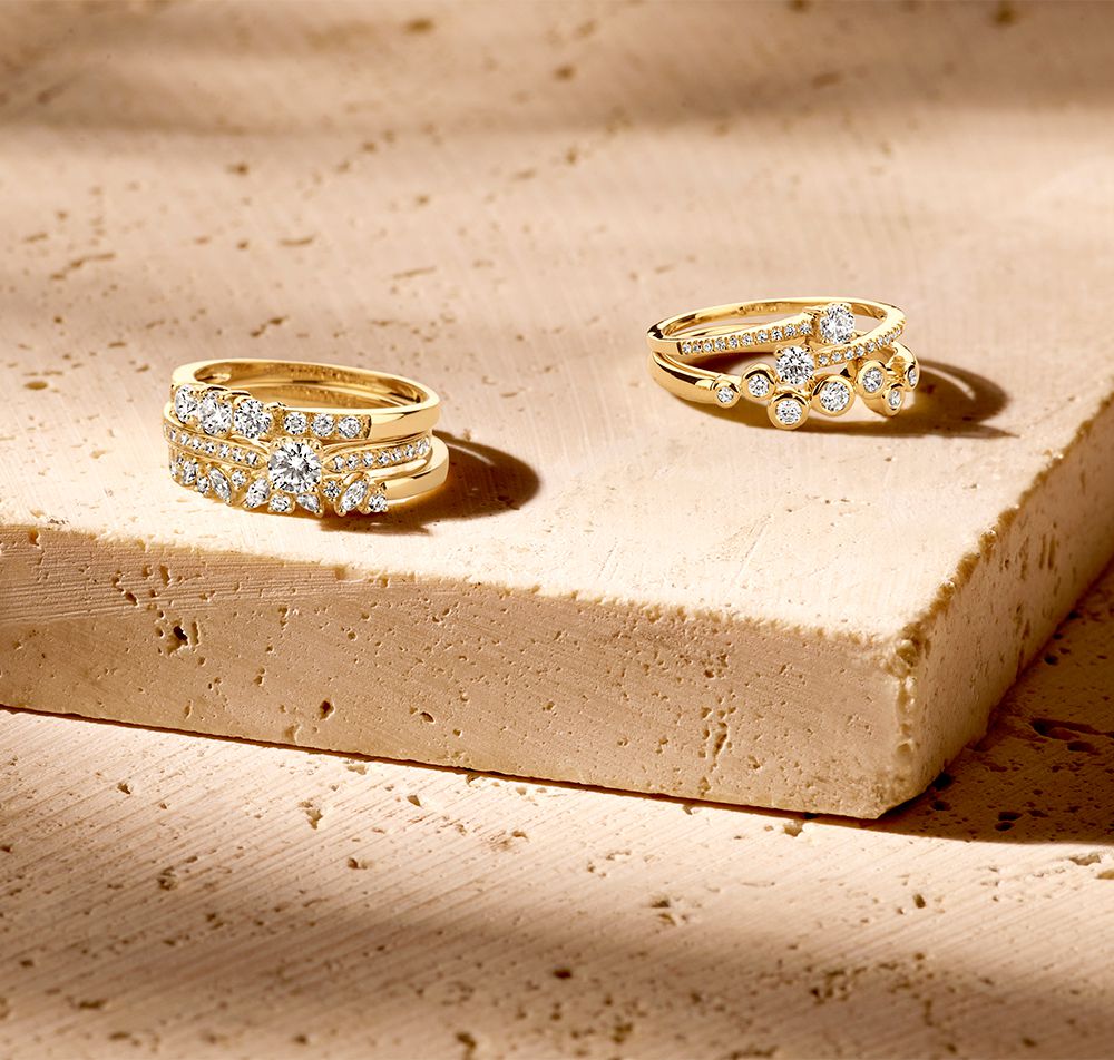 Twee 14 Karaat Geelgoud ringen van Excellent Jewelry, met tijdloos en romantisch ontwerp en zirkonia hartzetting, schitteren op een beige stenen oppervlak terwijl het zonlicht hun elegante schittering benadrukt.