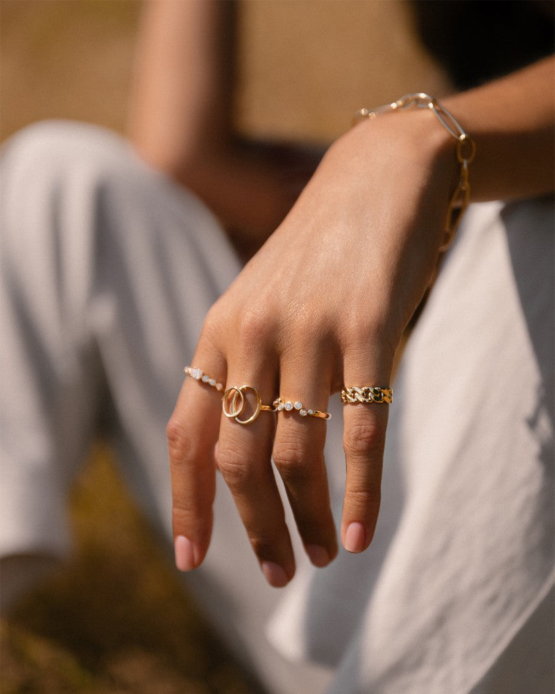 Een hand met een natuurlijke manicure draagt verschillende elegante gouden ringen, waaronder de Elegante Geelgouden Ring met Zirkonia's in Golvend Design - 14 Karaat Goud van Excellent Jewelry, plus een gouden armband. De persoon zit buiten in het zonlicht in een lichtgekleurde broek.