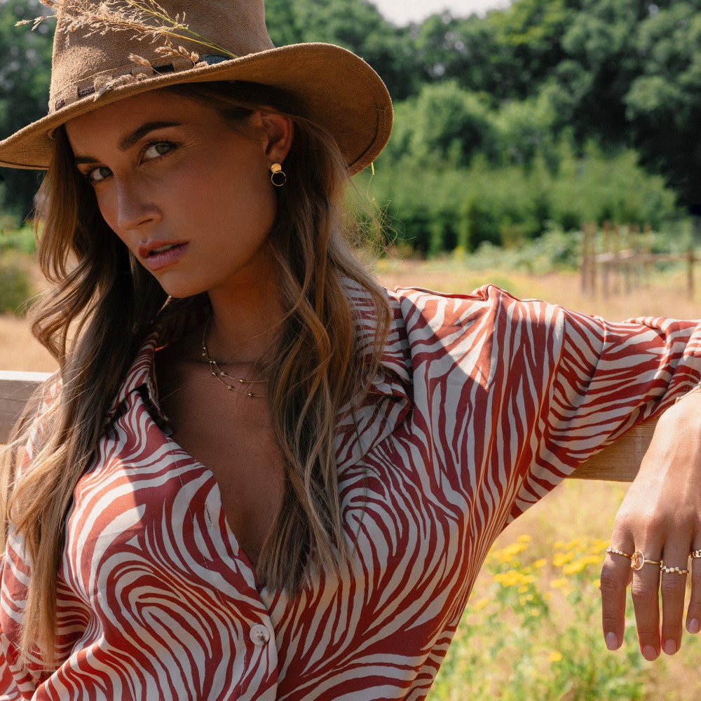 Een vrouw met een bruine hoed en rood-wit zebra-shirt leunt buiten op een houten hek en toont vol vertrouwen de Excellent Jewelry Elegante Geelgouden Ring met Zirkonia's in Golvend Design - 14 Karaat Goud. Groen vult de achtergrond.