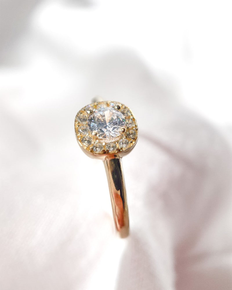 Een close-up van de Excellent Jewelry Geelgouden Ring met Ronde Zirkonia in Vierkante Zetkast en Halo - 14 karaat geelgoud, rustend op zacht lichtroze stof.