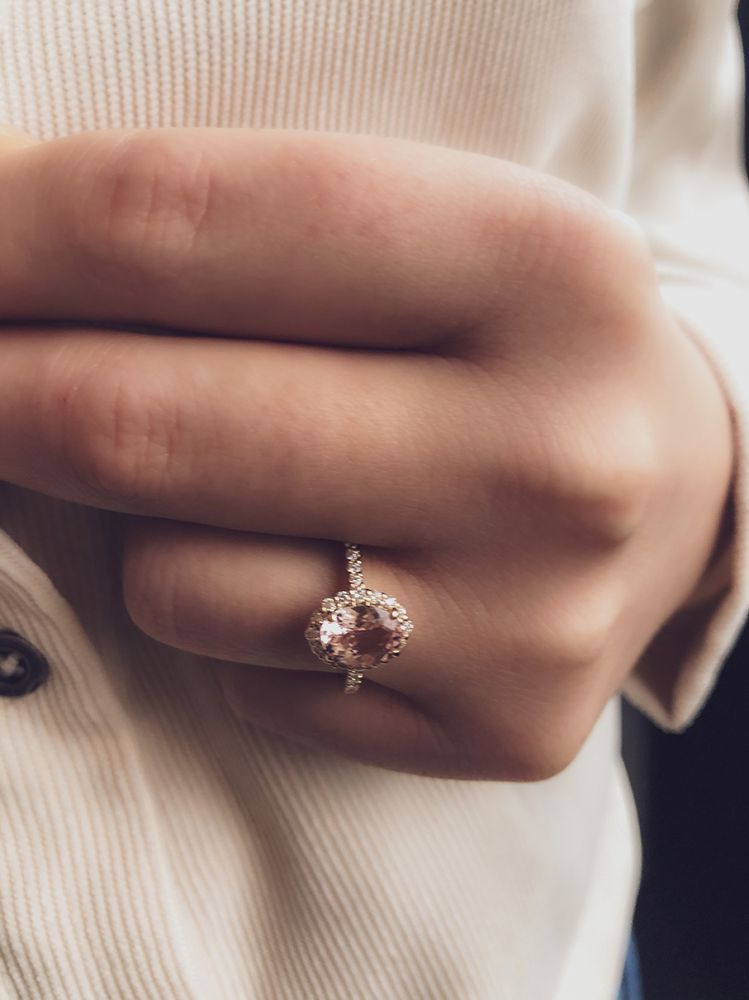 Een hand draagt de Excellent Jewelry 14 karaat roségouden ring met 1,28 ct ovale morganiet en 0,27 ct briljanten, met een grote ovale steen en diamanten in een dunne band. De persoon draagt een licht ribshirt.